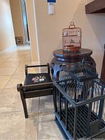 Photo shows two stools—a black wooden with floral embroidery and a round carved wooden stool—and two birdcages, one black metal and one bamboo-style.