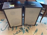 Two wood veneer bookshelf speakers with beige cloth grills standing side by side on a patterned rug