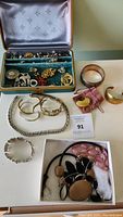 Photo showing jewelry box with assortment of brooches, earrings, and rings; three cuff bracelets; silver chain necklace; and large brooch with black and beige stones.