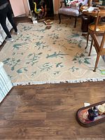 Full view of the large rectangular beige rug placed on wooden floor with blue and white floral patterns and white fringed edges, surrounding furniture partially visible.