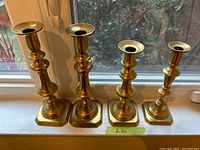 Four brass-tone metal candleholders displayed on a windowsill, showing varying heights and base styles; three hexagonal bases and one square base with circular tops for candles.