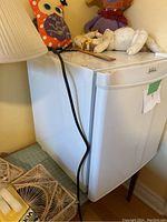 Closed view of the white Sunbeam mini fridge on a wooden stand with some stuffed toys on top, showing the power cord.