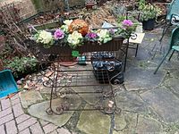Front view of rust-colored metal plant stand showing rectangular planter filled with ornamental gourds and flowers, with decorative scrollwork on legs and sides.
