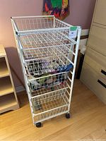 White metal rolling storage trolley with four wire basket drawers and black caster wheels. Front left angle shows basket construction and wheels.