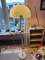 Standing floor lamp positioned near window with a large cream-colored plastic dome shade on a metal stand with round base.