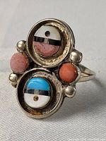 Front view of a sterling silver Zuni tribal ring showing two circular inlay designs with turquoise, coral, black, and white stones and additional round coral stones.