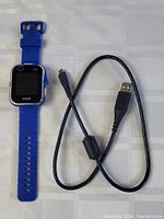 Blue VTech DX2 smart watch with visible VTech logo, orange and blue buttons on side, and black USB charging cable beside it on fabric background.