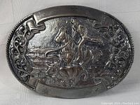 Front view of the silver-tone Western-themed belt buckle showcasing detailed embossed cowboy roping a calf while riding a horse, with a dog running alongside and decorative scrollwork on the border.