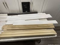 Photo showing assorted baseboards in natural wood, white painted, and a dark brown painted piece stacked on a table in front of white cabinets.