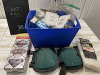 Blue plastic bin filled with assorted tools, packaging of nails, electrical outlet covers, plastic tubing, cheese cloth package, and two green knee pads in front of bin.