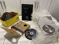 Lot overview with cardboard box containing metal cut off blades and multiple circular saw blades laid out on a white surface.