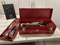 Open red metal tool box showing multiple hand tools inside including hammer, wrench, tape measure, etc.