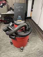 Photo showing the red vacuum cleaner with black hose attached, wheels visible on base, in a room with carpeted floor.