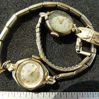 Pair of two gold filled gold plated vintage ladies watches placed on a black surface with sunlight highlighting metal finish and watch faces.