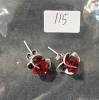 Photo of two silver earrings with red crystal gemstones lying on a black surface inside a plastic bag with a sticker '115'.