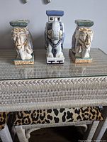 Front view of all three ceramic elephant stands arranged side by side on a glass tabletop showing the detailed painting and overall shape.