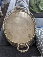 Top view of oval brass tray resting on fabric, showing interior with some tarnish and wear spots toward one end. The faux bamboo textured handles and rim are visible.