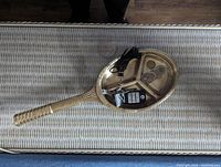 Top-down view of brass tennis racket laying flat on a woven white surface showing the hollow face and small decorative objects inside the racket head.