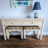Front view of white wicker console table with glass top, two drawers, and two leopard print cushioned stools below.