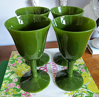Four green opaline water goblets arranged on a colorful surface, showing the full height and stem shape of the goblets clearly.