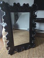 Front view of rectangular mirror showing ornate shiny black composite carved frame with leaf-like patterns.