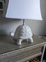 Full view of the white ceramic turtle lamp with a white rectangular lamp shade placed on a woven surface.