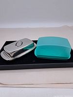 Two butter dishes side by side on black tray, metal and teal melamine dishes visible