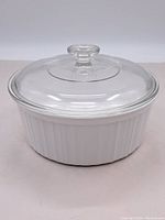 Front view of white oval ceramic casserole dish with clear glass lid and knob handle showing ribbed exterior detailing.