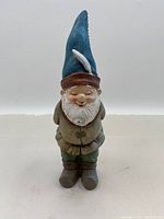 Front view of the garden gnome featuring blue pointed hat with white feather, white beard, beige and green clothing, and brown shoes.