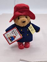 Front view of a Paddington Bear plush toy wearing red hat, blue coat, and red boots, holding a tag 'Classic Paddington Bear'