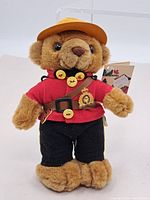 Front view of vintage RCMP Bear plush doll wearing Mountie uniform with red jacket, black pants, brown belt with gold buttons and yellow cap.