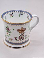 Side view of the mug showing the floral patterns, gold crown, 'R' emblem, and blue and gold trim.