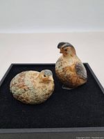 Two glazed ceramic stoneware grouse figurines displayed on a black surface, one standing and one in resting posture.