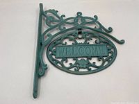 Full shot of the vintage cast iron welcome wall sign, showing bracket and hanging oval sign with ornate scrollwork in green paint with wear.