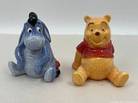 Front of the salt and pepper shakers showing Winnie the Pooh in yellow and red shirt, and Eeyore in blue-grey with a black mane, sitting.