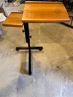 Side angle showing wooden adjustable table with larger main section and smaller side section, both mounted on black metal adjustable arms and rolling base.