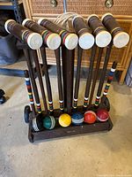 Photo showing the complete antique wooden croquet set in wooden rack with six mallets upright, six different colored balls lined up in front, and wickets stored in the rack.