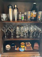 Wooden cabinet shelves displaying an assortment of barware including martini shakers, stemmed martini glasses, wine glasses, beer mugs, and decorative containers with coasters.