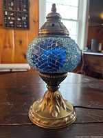 Small electric lamp with blue leaded glass shade and brass-colored metal base on wooden table with background of window and kitchen.