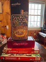 Stack of board games and puzzle including Mordillo Crazy Circus 1500-piece puzzle, Zumba Exhilarate DVD, Scrabble and Monopoly games.