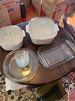 Overview photo showing collection of white and clear Pyrex dishes including square cookware dishes with lids, rectangular glass baking dish, and stack of small clear dessert dishes.
