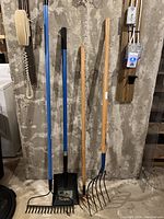 Photo showing blue-handled bow rake, digging shovel with squared blade, and wood-handled cultivator rake with curved tines leaning against concrete wall