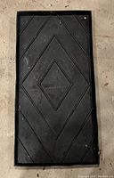 Flat rectangular black boot rack made of heavy-gauge rubber. Features an embossed geometric diamond and diagonal line pattern for traction and water containment. Slight wear visible from light use.