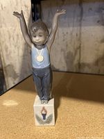 Front view of porcelain figurine of child standing on a base decorated with Olympic torch and leaves, gold medal visible