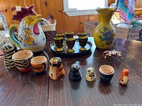 Photo showing the full lot on wooden table with ceramic roosters and flower pitchers, wooden cat nesting dolls, Russian hand-painted shot glasses on a tray.