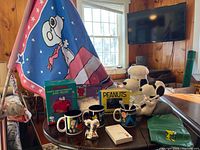 Photo showing full collection on a round table including Snoopy porch flag, coffee mugs, paperweight and birdfeeder box