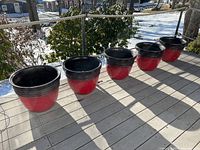 Five red and black resin flower pots stacked next to each other outdoors on a wooden deck with snow in background.