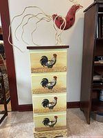 Wooden cabinet with four drawers, each drawer front painted with a black, red, and white rooster on a beige background, standing on floor next to wall.