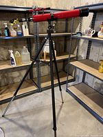 Full view of the red Tasco telescope mounted on a black adjustable tripod stand in a basement storage space.