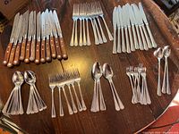 Photo showing complete set of 12 Oxford Hill steak knives with wooden handles arranged in the center with multiple Towle stainless steel forks, knives and spoons grouped around them on wooden table.